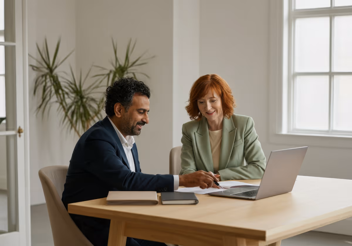 Two business professionals sitting at a desk reviewing documents with a laptop in a bright office.