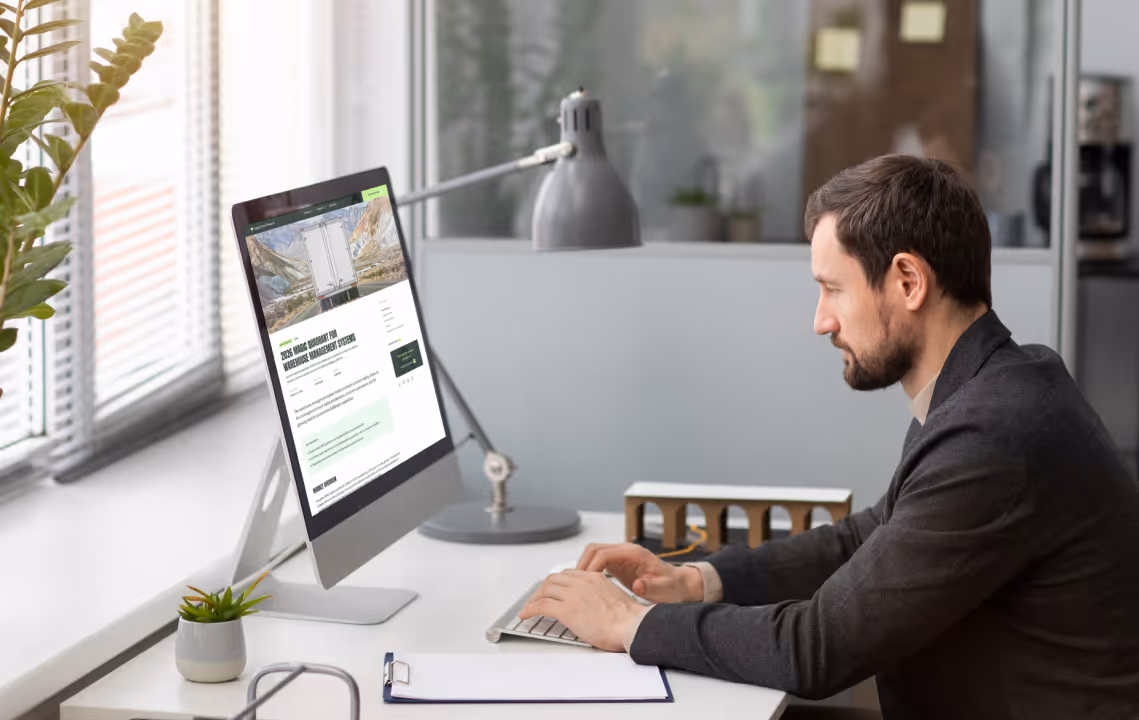 Man in a dark blazer working on a desktop computer at a bright office desk with plants and a lamp.