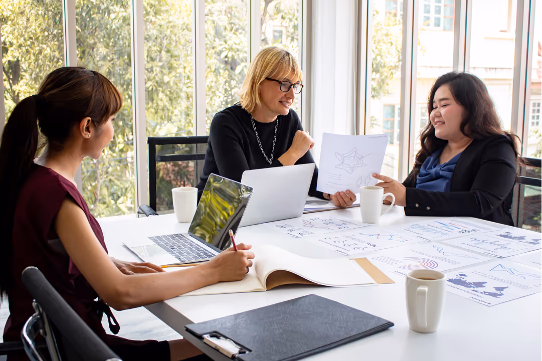 Three women in a bright office discussing charts and documents around a table with laptops and coffee cups.