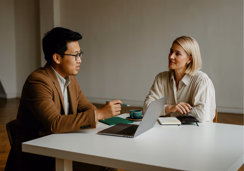 Two colleagues, a man and a woman, having a discussion at a white table with a laptop and notebooks.