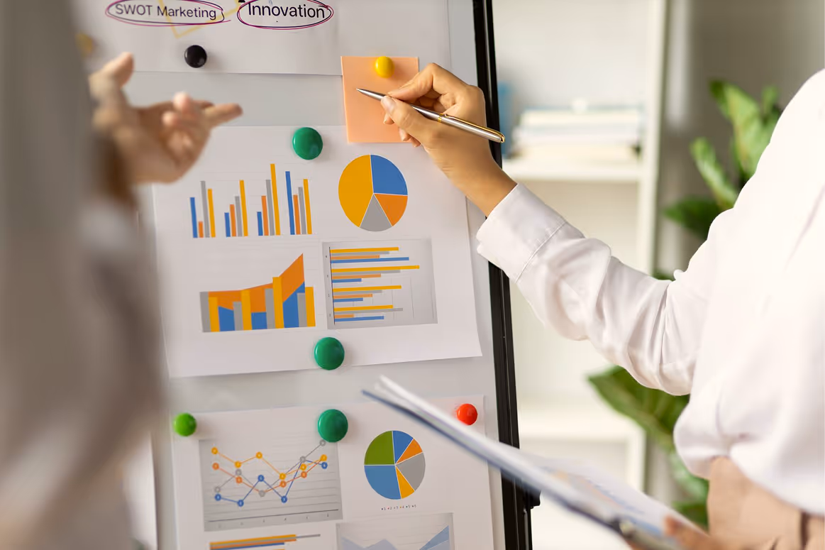 Two people discussing colorful charts and graphs on a whiteboard with one holding a pen and sticky note for planning.